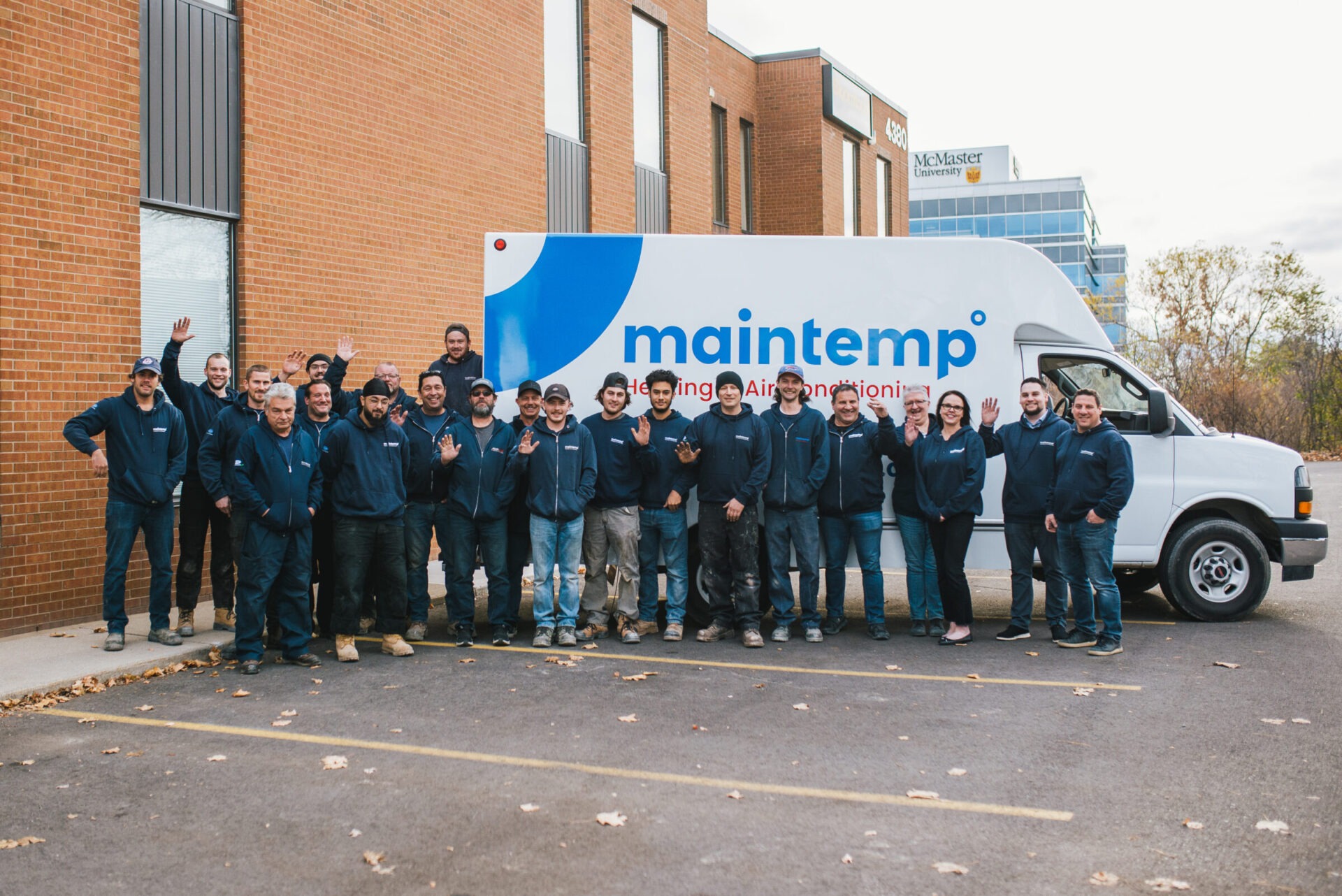 A group of smiling people in matching work uniforms pose in front of a large van with "maintemp" logo, outside a brick building with trees.