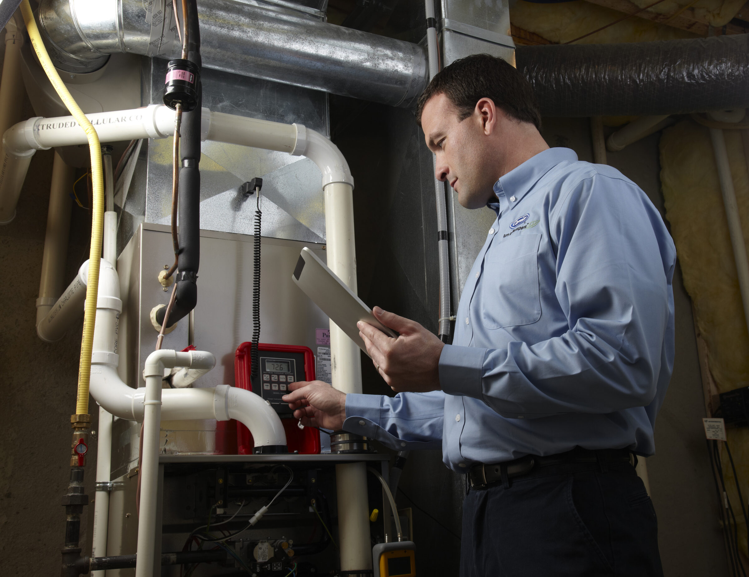 A person in a blue shirt is inspecting a heating system, using a tablet, with white PVC pipes and insulation in the background.