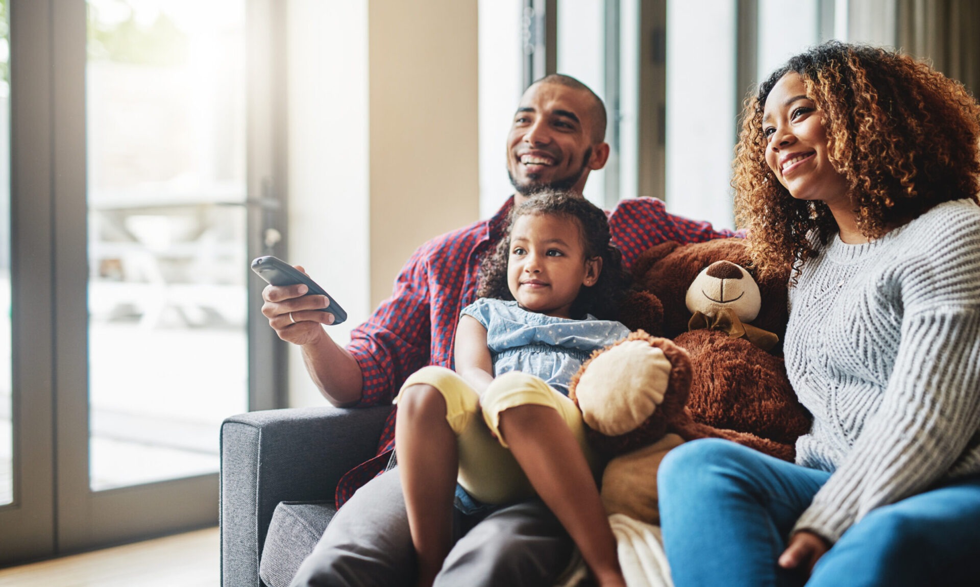 A family of three, two adults and a child, are sitting together comfortably with a teddy bear, while the person holds a remote, smiling.